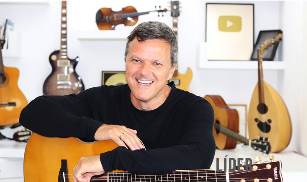 Uma foto de Heitor Castro, homem branco sorrindo, camisa preta de mangas longas, anel dourado no dedo anelar, na mão direita, sentado, em primeiro plano, com um violão. Heitor está com os braços cruzados em cima do violão. Ao fundo, um pouco desfocado, estão vários instrumentos de corda, como outro violão, uma guitarra elétrica, um violino, possivelmente outra guitarra, outro violão deitado, e um alaúde. Também há destaque para as placas do YouTube, a dourada e a prata.